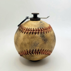 Könnte beinhalten: Ein baseballförmiger Flaschenverschluss mit einem gealterten, hellbraunen Leder-Look. Die roten Nähte sind deutlich sichtbar, und der Verschluss hat eine dunkelbraune Oberseite mit einem silbernen Metallgriff und einem schwarzen Ring.