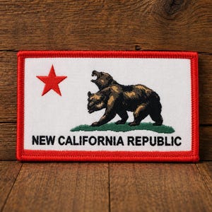 May include: A rectangular embroidered patch featuring a white background with a red border. It displays a red star on the left, a brown bear standing on a green patch of grass in the center, and the black text NEW CALIFORNIA REPUBLIC below.