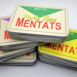 Puede incluir: Una pila de latas rectangulares de metal con esquinas redondeadas. Cada lata tiene un fondo color crema con la palabra "MENTATS" en rojo y "Party Time" en letras doradas. Las latas tienen un triángulo verde o amarillo.