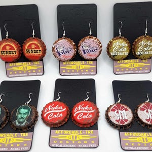 Könnte beinhalten: Ein Set mit 10 Paar Ohrringen aus Flaschenverschlüssen. Die Flaschenverschlüsse zeigen verschiedene Designs, darunter die Worte "Sunset Sarsaparilla", "Vim", "Nuka Cola" und "SM". Die Ohrringe sind hypoallergen und nickelfrei.