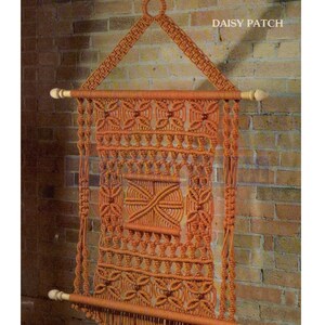May include: A macrame wall hanging with a geometric pattern in shades of orange. The hanging is made with thick cord and has a wooden dowel at the top and bottom. The hanging is labeled "Daisy Patch".