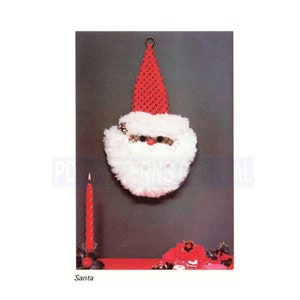 May include: A red and white Santa Claus head made of yarn with a red hat and a white beard. The Santa head is hanging on a wall with a red candle in front of it.