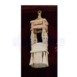 Può includere: Un'appendice in macramè bianca con una base quadrata e un'apertura rettangolare. Il macramè ha una frangia sul fondo ed è decorato con accenti rossi e bianchi.