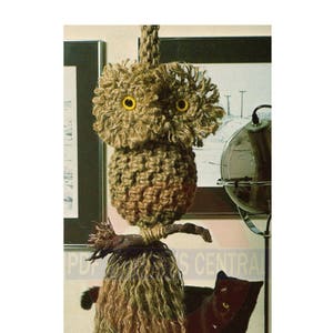 Puede incluir: Un búho marrón y beige hecho de cuerda con ojos amarillos colgando del techo. Un gato negro está de pie frente al búho.