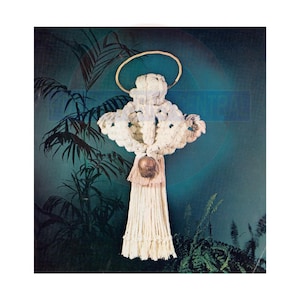 Op de afbeelding: Een witte macramé engel met een gouden halo en een bruine bel die eronder hangt. De engel hangt tegen een groene achtergrond.