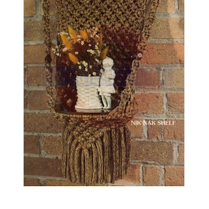May include: A brown macrame wall shelf with a woven basket and a small figurine on top. The shelf is hanging on a brick wall and has the text "Nik Nak Shelf" printed below it.