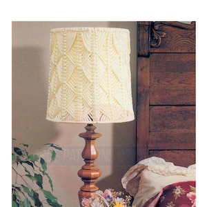 May include: A white crocheted lampshade with a geometric pattern sits on a wooden lamp base. The lamp is on a bedside table next to a bed with a floral patterned quilt and a book.