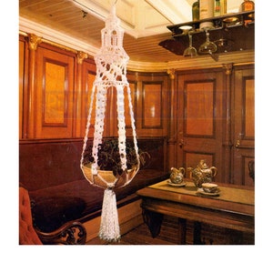 May include: White macrame plant hanger with a woven basket and a tassel hanging from the bottom. The hanger is suspended from a hook in a room with wood paneling and a table with a teapot and cups.