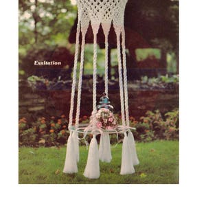 May include: A white macrame hanging plant holder with a round glass shelf and white tassels. The plant holder is hanging in a garden setting with green grass and flowers.