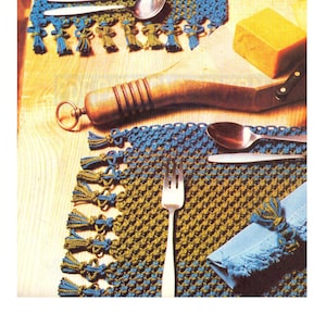 May include: Two crocheted placemats with blue and green fringe. The placemats are made with a knotting technique and have a diamond pattern. A blue napkin is rolled up on one placemat and tied with a knot.
