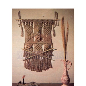 May include: A brown macrame wall hanging with a wooden dowel at the top. The hanging has a geometric pattern and is decorated with tassels. A pink ceramic vase with a floral design is on a wooden table next to a small candle.