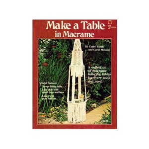 Puede incluir: Portada de libro con una mesa colgante de macramé blanca en un entorno de jardín. El título del libro es "Make a Table in Macrame" y los autores son Cathy Bonds y Carol Millsaps. La portada del libro presenta una colección de mesas colgantes de macramé para cada rincón y necesidad.