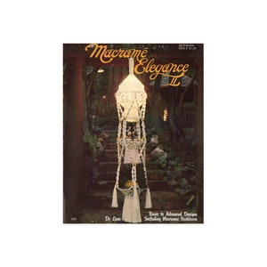 May include: A book titled "Macrame Elegance II" with a white macrame hanging in the background. The book cover features a photo of a white macrame hanging in a garden setting. The book cover also includes the text "Basic to Advanced Designs Including Macrame Necklaces".