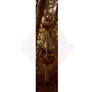 Puede incluir: Un macetero de macramé con dos macetas marrones, una con una planta verde, colgando de una rama de árbol en un entorno boscoso. El texto "La Belle" es visible en la parte inferior de la imagen.