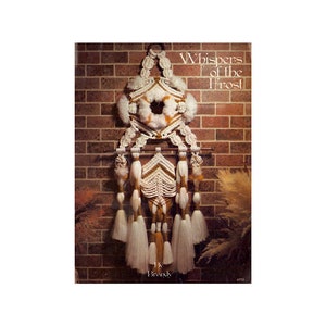 Op de afbeelding: Een macramé wandhanger in wit en bruin. De hanger heeft een grote cirkel in het midden en een reeks kwastjes aan de onderkant. De titel "Whispers of the Frost" staat op de afbeelding gedrukt.
