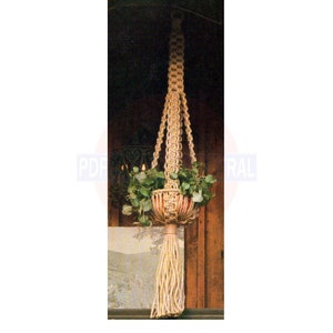 Puede incluir: Un macetero de macramé de color beige natural, que sostiene una pequeña maceta de terracota con plantas verdes. El macetero tiene un borlón en la parte inferior.