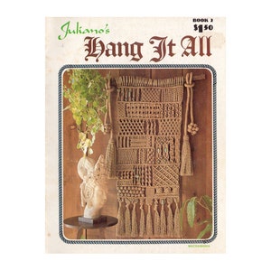 Pode incluir: Uma tapeçaria de macramé de parede com um esquema de cores castanho e bege. A tapeçaria apresenta uma variedade de nós e padrões, incluindo franjas e contas. O título "Juliano's Hang It All" e "Book 2 $1.50" estão impressos na página.