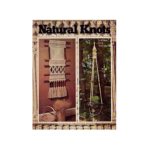 Può includere: Una copertina di libro vintage intitolata "Natural Knots" con portavasi in macramè e arazzi da parete. L'immagine mostra esempi di design in macramè, tra cui un arazzo da parete e un portavaso sospeso. La copertina ha un'estetica retrò.