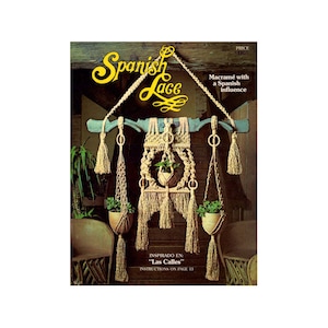 Puede incluir: Una portada de libro con el título "Spanish Lace" en letra amarilla. La portada presenta una pared de macramé con tres colgadores de plantas, inspirado en "Las Calles".
