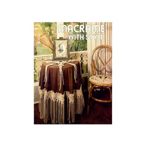 May include: A brown table with a white macrame tablecloth and fringe. A framed picture sits on the table. A wooden chair with a macrame seat sits next to the table. The text "Macrame' with Style" is printed above the table.