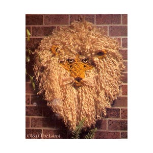 Pode incluir: Um leão de crochê pendurado na parede com um fundo de tijolo marrom. O leão tem uma juba fofa e um rosto amarelo com olhos pretos.