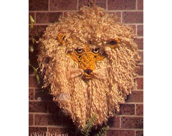 May include: A crocheted lion head wall hanging with a brown brick background. The lion has a fluffy mane and a yellow face with black eyes.