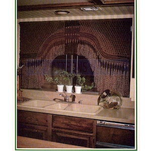 May include: A brown crocheted window valance with fringe hangs over a kitchen sink with a double basin. The sink is made of white porcelain and has a chrome faucet. There are three potted plants on the windowsill behind the sink.
