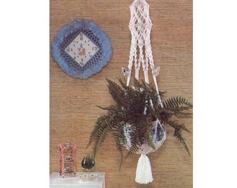 May include: White macrame plant hanger with a white tassel hanging from the bottom. A green fern plant is in the pot. The hanger is hanging from a ceiling against a brown wall.