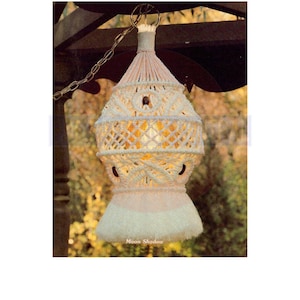 Op de afbeelding: Een witte macramé lampenkap met een gloeilamp erin, hangend aan een ketting. De lampenkap heeft de vorm van een traan en heeft een franje aan de onderkant. De tekst "Moon Shadow" is zichtbaar onder de lampenkap.