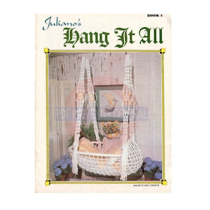 May include: A white macrame cradle hanging from the ceiling. The cradle is made of a thick rope and has a white fabric lining. The cradle is decorated with white flowers and a white blanket. The text "Snowflake Cradle" is printed below the cradle.