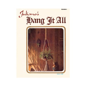 Può includere: Una copertina di libro con il titolo "Juliano's Hang It All" in lettere rosse. La copertina presenta una foto di un portavasi in macramè con una pianta verde al suo interno. Il portavasi è appeso a un soffitto di legno.
