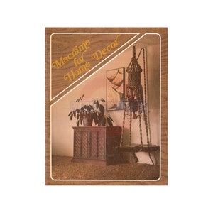 Op de afbeelding: Een boek getiteld "Macrame voor Home Decor" met een bruine houtnerfomslag. De omslag toont een macrame hangende plantenhouder in een woonkamer.