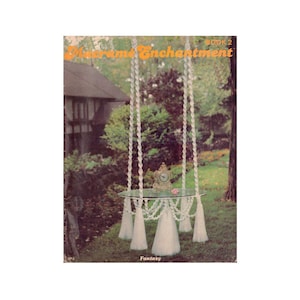 Peut inclure: Une table suspendue en macramé blanche avec un plateau en verre et des glands blancs. La table est suspendue à une branche d'arbre dans un jardin. La couverture du livre indique "Macramé Enchantment Livre 2" et "Fantasy".