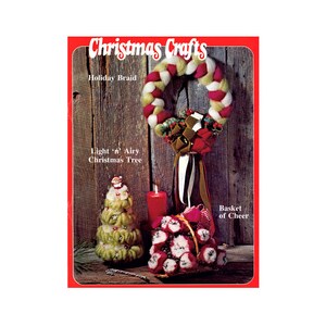 May include: A festive image featuring handmade Christmas crafts. A braided wreath with red, white, and green yarn hangs above a basket of red and white ornaments. A small, green Christmas tree and a red candle complete the holiday scene. The text "Christmas Crafts" is at the top.
