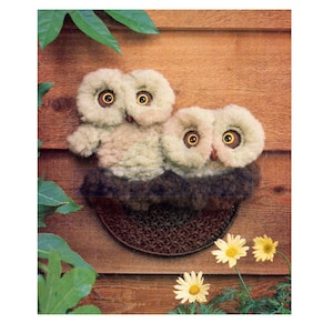 Puede incluir: Dos búhos de crochet con ojos marrones y pelaje beige y marrón sentados en un nido de crochet marrón. Los búhos están sobre un fondo de madera con hojas verdes y flores amarillas.