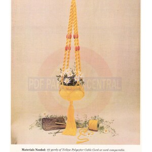 May include: A yellow macrame plant hanger with eight orange beads and a yellow tassel. The hanger is holding a yellow pot with white flowers. Materials needed: 66 yards of yellow polyester cable cord or cord comparable, eight orange beads, one 2 1/2 inch solid metal ring.