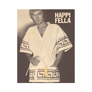 May include: A black and white photograph of a person wearing a white robe with a black and white geometric pattern. The robe has a belt and the person is holding the belt in their hand. The text "HAPPI FELLA" is printed in the top right corner of the photograph.