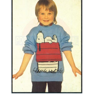 May include: A blue knitted sweater with a white and black image of Snoopy lying in his red doghouse. The sweater is worn by a young child with blonde hair.