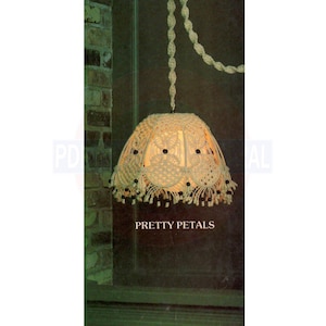 May include: A macrame lampshade with a white cord hanging from a brick wall. The lampshade is decorated with black beads and has a label that reads "Pretty Petals".