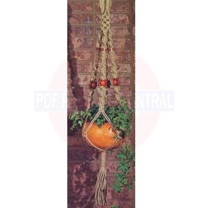 May include: A macrame plant hanger with a woven design, holding a round orange pot filled with green plants. The hanger is made of natural-colored rope and features decorative wooden beads. The background is a brick wall.
