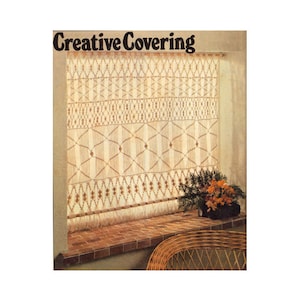 Op de afbeelding: Een close-up van een raam met een macramé gordijn. Het gordijn is gemaakt van natuurlijke vezels en heeft een franje aan de onderkant. Het gordijn hangt voor een raam met een bakstenen vensterbank. Er staan bloemen in een pot op de vensterbank. De tekst "Creative Covering" staat bovenaan de afbeelding.