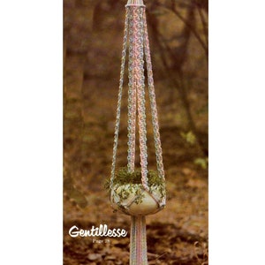Puede incluir: Un macramé para colgar plantas con una maceta blanca y una planta verde. El macramé está hecho con hilo rosa, azul y blanco y tiene borlas largas. El texto "Gentillesse Página 28" es visible en la parte inferior de la imagen.