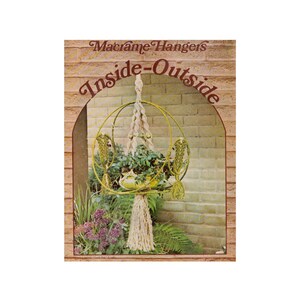 May include: A macrame plant hanger with a round, woven design and a long tassel. The hanger is hanging from a brick wall and has a potted plant inside.
