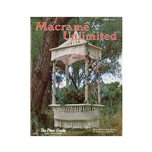 May include: A white macrame plant cradle hanging from a tree branch. The cradle is shaped like a gazebo with a fringed roof and a round base. The text "The Plant Cradle" and "Page Twenty-Three" is visible at the bottom of the image.