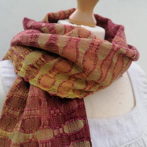 May include: A red, orange and yellow patterned woven scarf with a geometric design. The scarf is draped around a white mannequin.