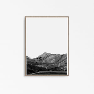 May include: Black and white framed print featuring a mountain landscape. The photograph captures the texture and shadows of the mountains, with a bright white sky above. The frame is a light wood color, adding a natural touch to the artwork.