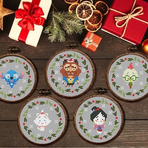 Peut inclure: Cinq cercles de broderie au point de croix avec des personnages de dessins animés, dont Stitch et La Bête, sont présentés sur une surface en bois. Chaque motif est encadré par une couronne verte et rouge. Des cadeaux et des ornements de Noël entourent les cercles.
