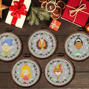 May include: Five cross-stitch ornaments in wooden hoops, each featuring a cartoon character with a Christmas theme. The characters include a dog, a fox, and three other figures, all surrounded by a green and white wreath on a gray background.