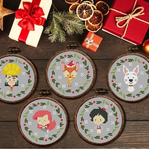 May include: Five round cross-stitch ornaments with cartoon characters, each framed in a wooden hoop. The characters include a girl with a yellow hat, a fox in a Santa hat, a white dog, a girl with red hair, and a girl with a black bow. The background is gray.