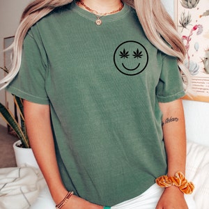 May include: Green short-sleeved t-shirt with a black smiley face design. The eyes of the smiley face are cannabis leaves. The shirt is a solid green color.